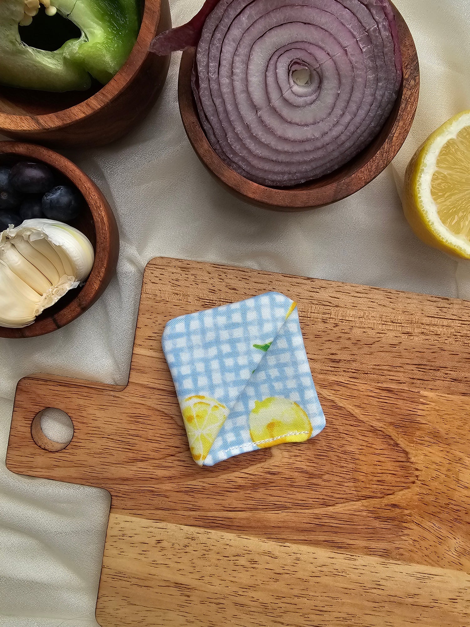 A white and blue plaid corner bookmark with a design of lemons placed on a wooden cutting board, surrounded by various kitchen items like garlic, lemon, and sliced onion.