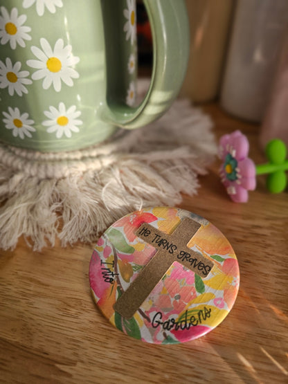 Round pin button with floral design and text on a cross that reads, "He turns graves into gardens". Button is on a wooden surface next to a green container with white flowers.