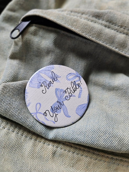 Round pin button with blue bows on white background and lettering that reads, "Read Your Bible!". Button sits on a blue denim bag.
