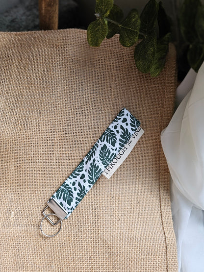 A fabric keychain strap with a white background and dark green tropical leaf pattern, featuring a silver metal key ring, displayed on a burlap surface.