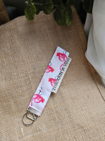 A close-up of a fabric keychain strap with a silver metal split ring, featuring a white background with pink flamingo print. The strap is resting on a textured beige burlap fabric background.
