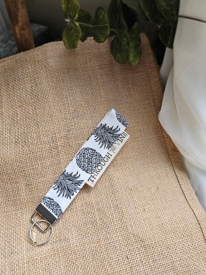 A fabric keychain strap with a white background and black pineapple pattern, resting on a burlap surface.