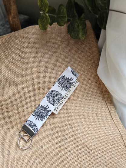 A fabric keychain strap with a white background and black pineapple pattern, resting on a burlap surface.