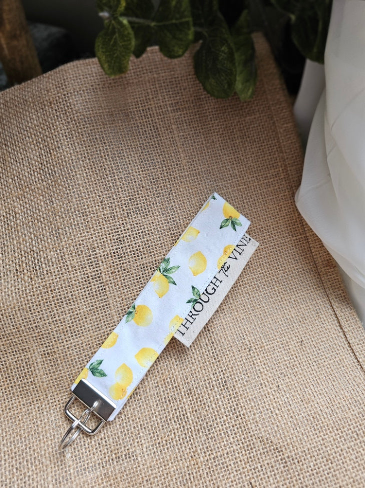 A fabric keychain or bag clip with a white background, yellow lemon print, and green leaves, resting on burlap.