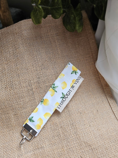 A fabric keychain or bag clip with a white background, yellow lemon print, and green leaves, resting on burlap.