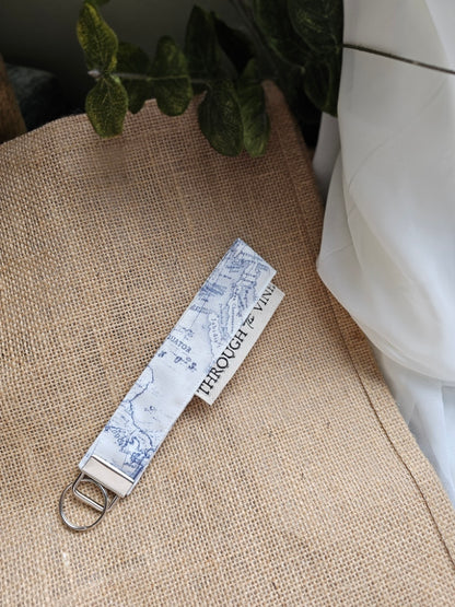 A fabric keychain or bag strap with a blue map print on white, attached to a silver metal ring, resting on a beige burlap surface with a white fabric and green leaves in the background.