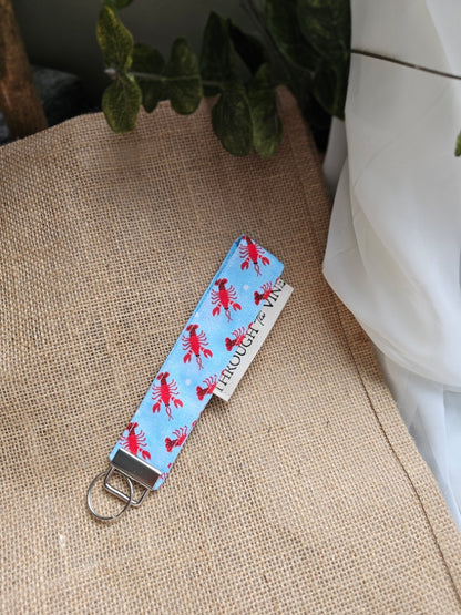 A fabric wristlet keychain strap with a light blue background and a repeating red lobster print, resting on burlap.