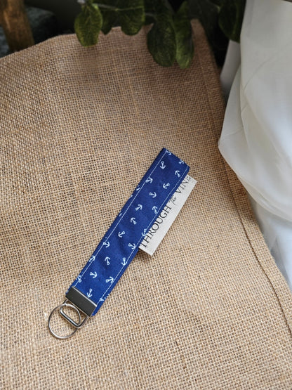 A fabric wristlet strap with a blue and white anchor pattern, featuring a silver metal D-ring, displayed on a beige burlap surface.