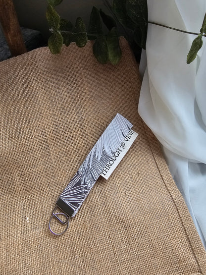 A fabric wristlet strap with a brown and white palm leaf pattern and a silver metal key ring, resting on a burlap surface next to white fabric and green leaves.