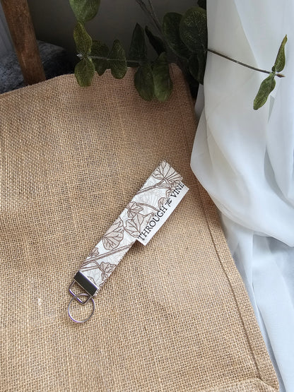 A fabric keychain strap with a botanical line pattern on a white background, featuring a silver metal clasp and keyring, resting on a burlap surface with white fabric and green leaves in the background.