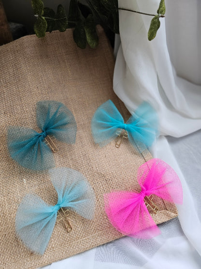 Four tulle bookmarks in pink, light and medium blue, and teal, arranged on a burlap fabric background with white draped fabric and green leaves as props.