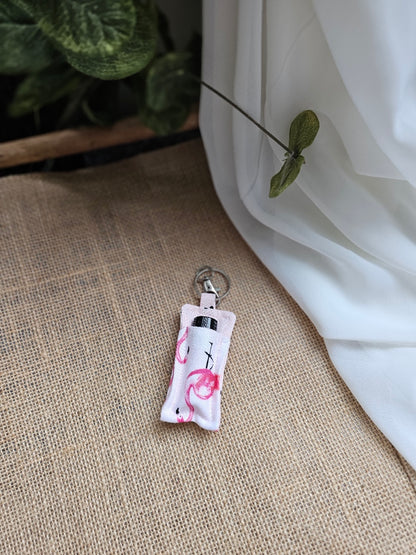 A small fabric keychain oil keeper with flamingo pattern, resting on a textured beige burlap surface next to a white drape of fabric and green leaves.