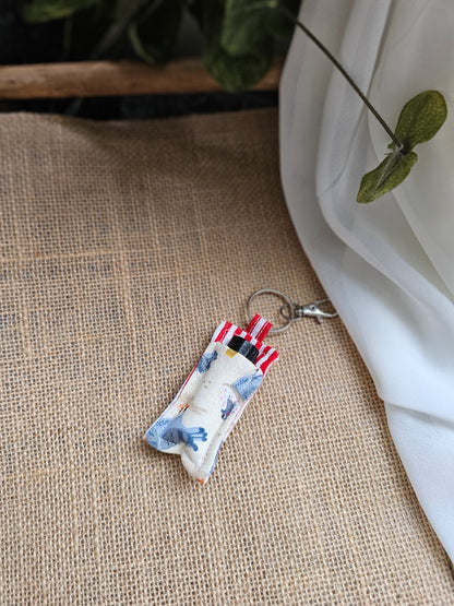 Oil Keeper (striped red/white on pigeon fabric background) resting on a beige burlap surface with white fabric and green leaves in the background.