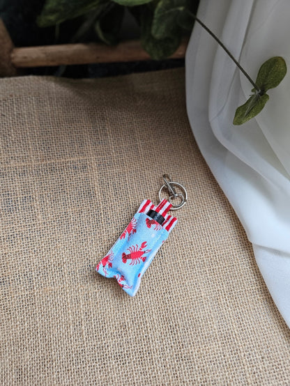 A small fabric keychain oil keeper with a lobster print. It is displayed on a textured beige burlap surface with a white fabric drape and green leaves in the background.
