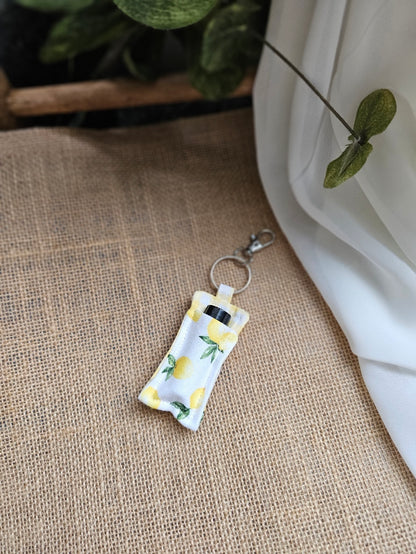 A small fabric keychain oil keeper with lemon fabric, resting on a textured beige burlap surface with white fabric and green leaves in the background.