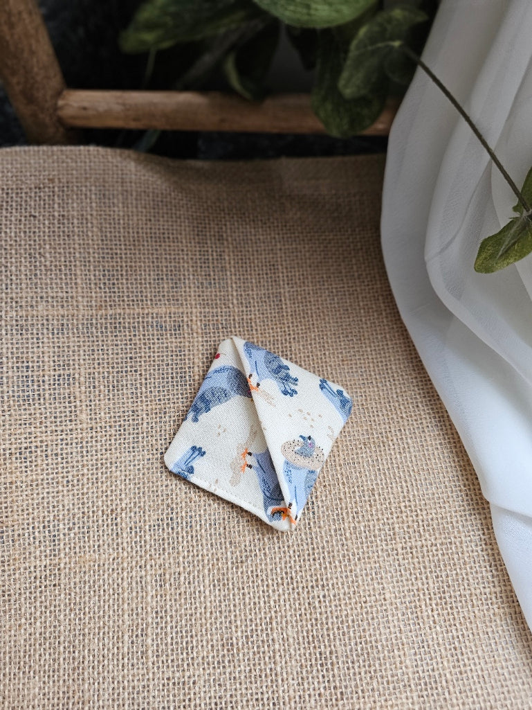 A small folded square fabric corner marker with pigeons on light background patterned print rests on a beige burlap surface next to a white draped fabric and green leaves.