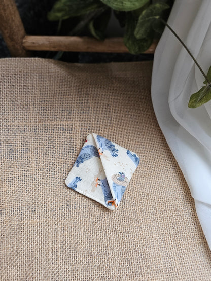 A small folded square fabric corner marker with pigeons on light background patterned print rests on a beige burlap surface next to a white draped fabric and green leaves.