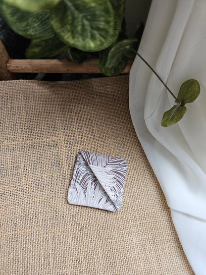 A small square folded fabric corner marker with a brown and white leaf pattern rests on a beige burlap surface, with white draped fabric and green leaves in the background.