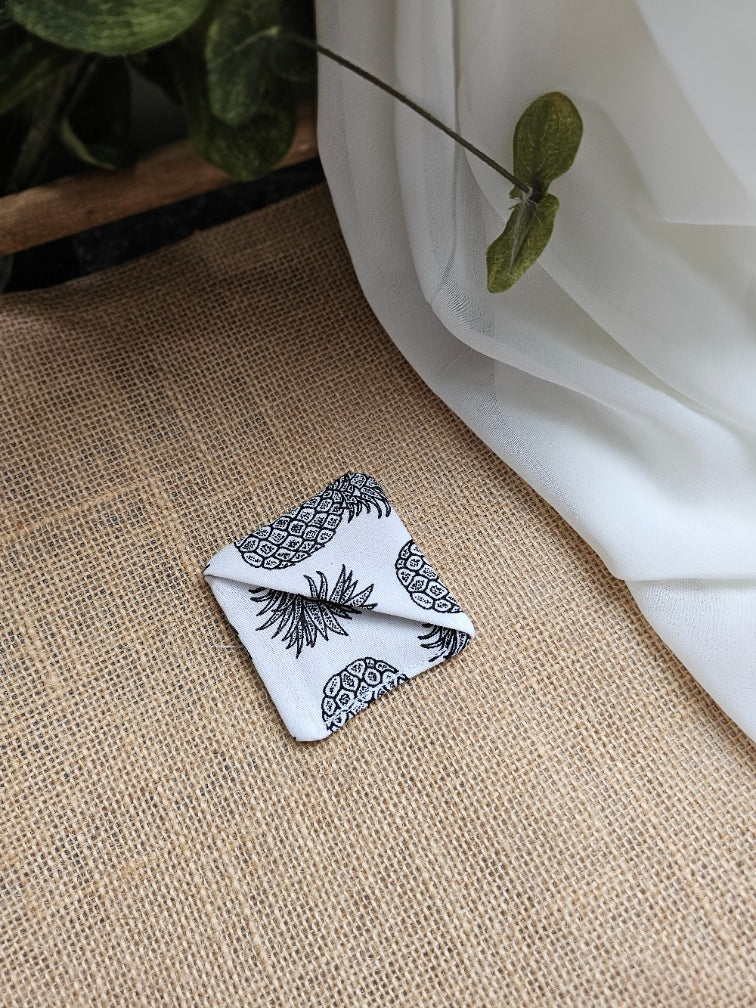 A small folded white fabric corner marker with a black pineapple print pattern, placed on a beige woven surface with white draped fabric and green leaves in the background.