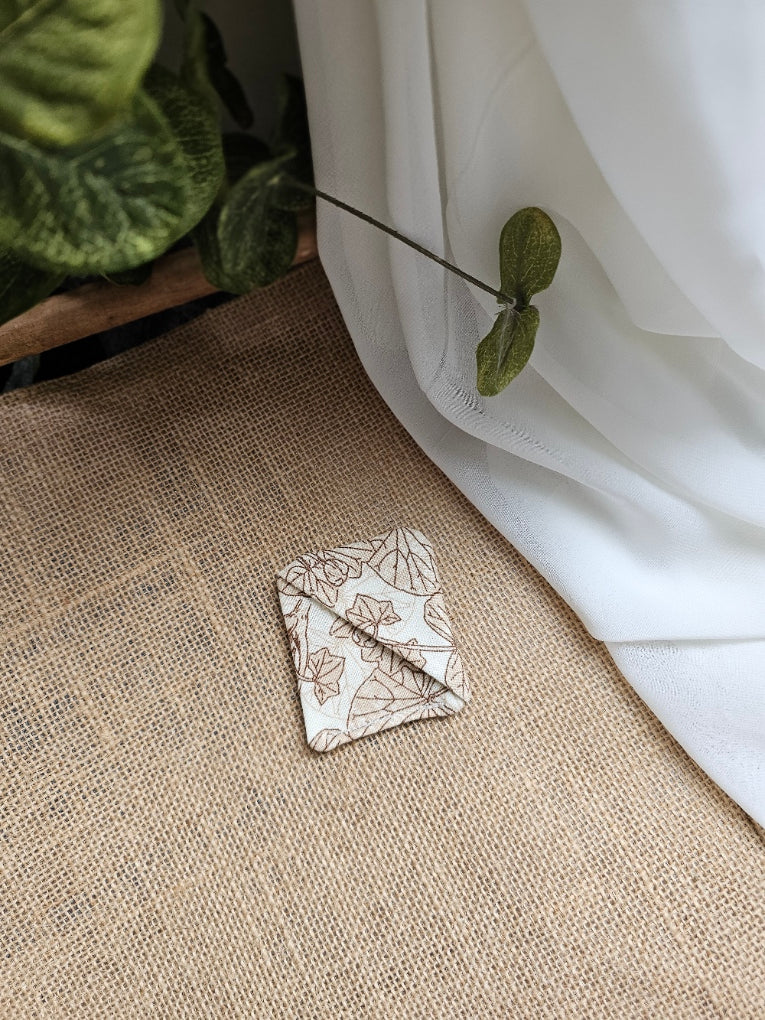A small square fabric corner marker with a beige and brown floral pattern, resting on a textured beige burlap surface next to a white drape of fabric and green leaves.