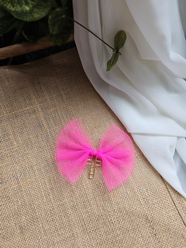 A bright pink tulle bow bookmark with a gold metal cross, displayed on a textured beige fabric surface next to a draped white fabric and green leaves.