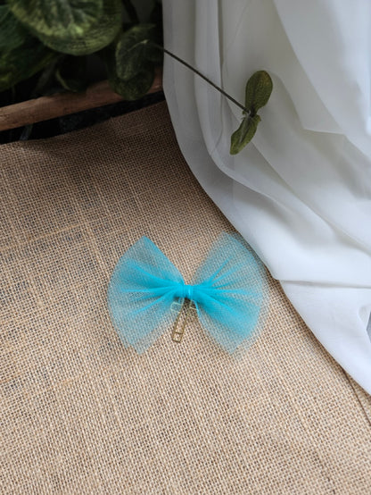A light blue tulle bookmark with a small gold metal cross, resting on a textured beige burlap surface next to a white fabric drape.