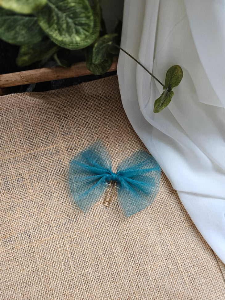 A dark blue tulle bookmark with a gold metal cross clip, displayed on a beige burlap fabric surface next to a white drape and green leaves.