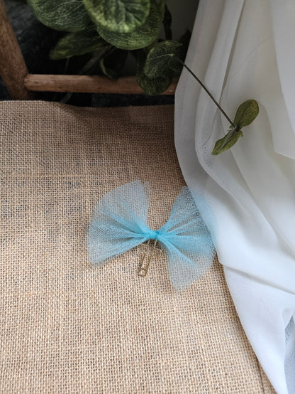 A light blue tulle bookmark with a cross, resting on a beige burlap fabric surface next to white draped fabric and green leaves.