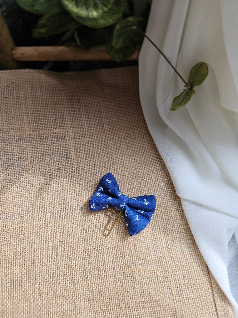 A small blue fabric bow with a white anchor pattern, resting on a textured beige burlap surface with white fabric and green leaves in the background.