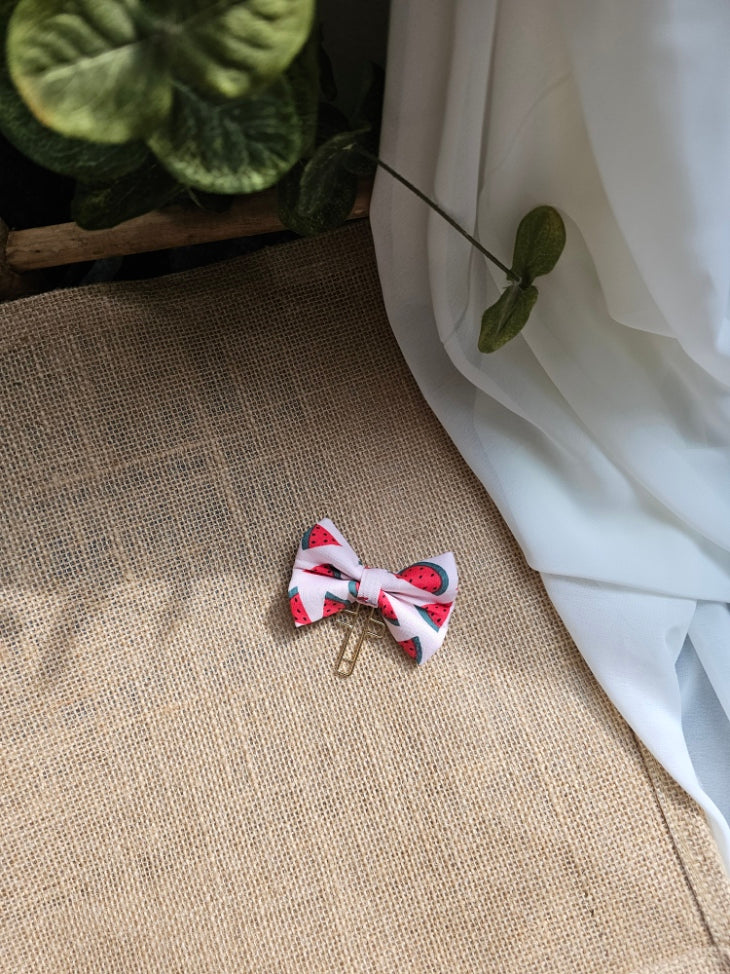 A small fabric bow bookmark with a red and green watermelon print and a gold metal clip, resting on a burlap surface next to white fabric and green leaves.