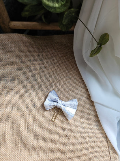 A small fabric bow with a white and blue  map pattern, attached to a gold-tone metal cross, resting on a textured burlap surface with white fabric and green leaves in the background.