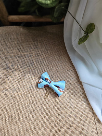 A small fabric bow bookmark with a coconut on light blue background pattern, attached to a gold-tone cross, resting on a textured beige burlap surface.