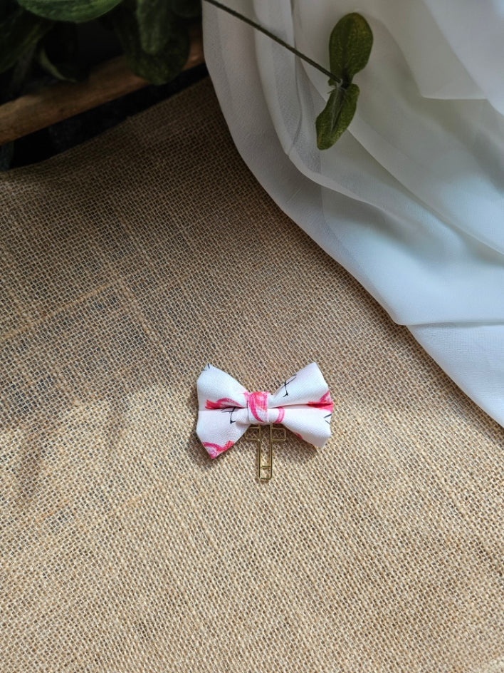 A small fabric bow bookmark, with a flamingo pattern, attached to a gold-tone cross. It is placed on a textured beige burlap surface with a white fabric drape and a green plant stem in the background.