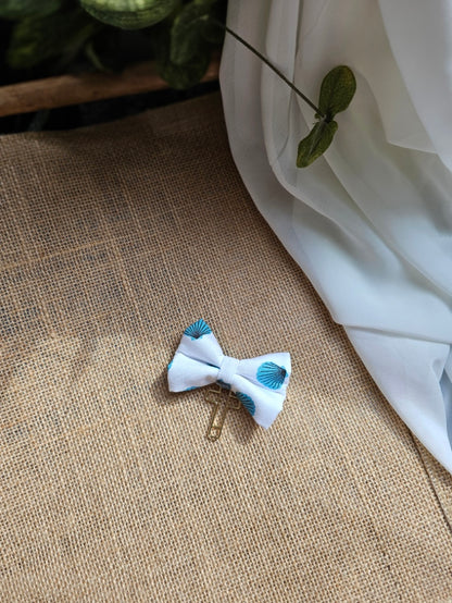 A small fabric bow bookmark with a seashell pattern, resting on a brown burlap surface with white fabric and green leaves in the background.
