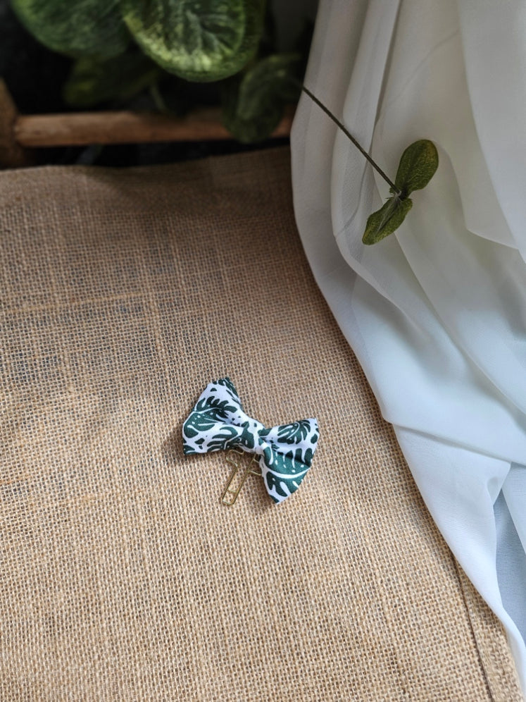 A small fabric bow bookmark with a palm patterned design, resting on a textured burlap surface with draped white fabric and green leaves in the background.