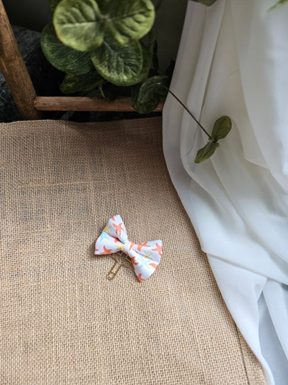 A small fabric bow bookmark with a starfish print, resting on a textured burlap surface next to a white drape and green leaves.