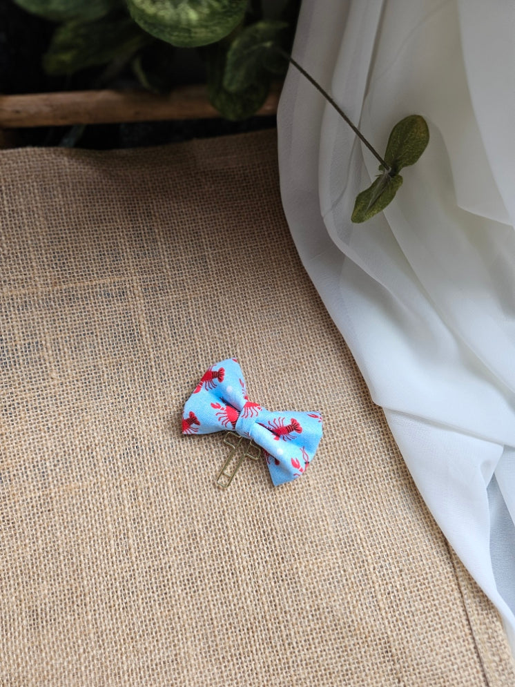A small fabric bow bookmark with a lobster print, resting on a textured beige burlap surface with white fabric and green leaves in the background.