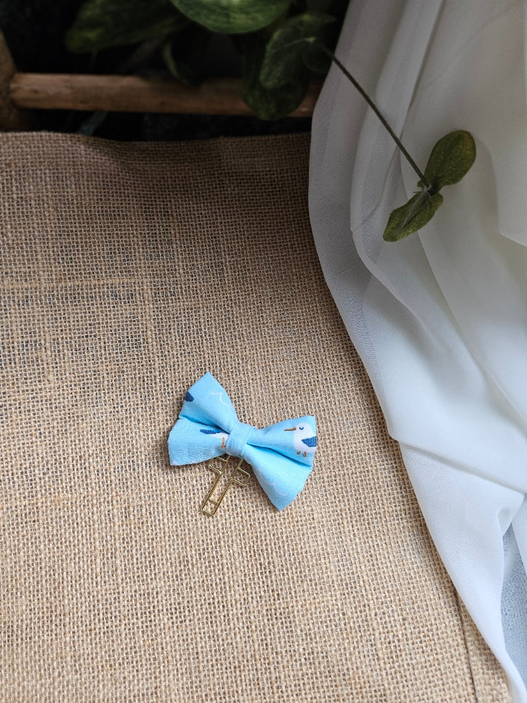 A small light blue fabric bow bookmark with a pigeon design, resting on a textured beige burlap surface with white draped fabric and green leaves in the background.