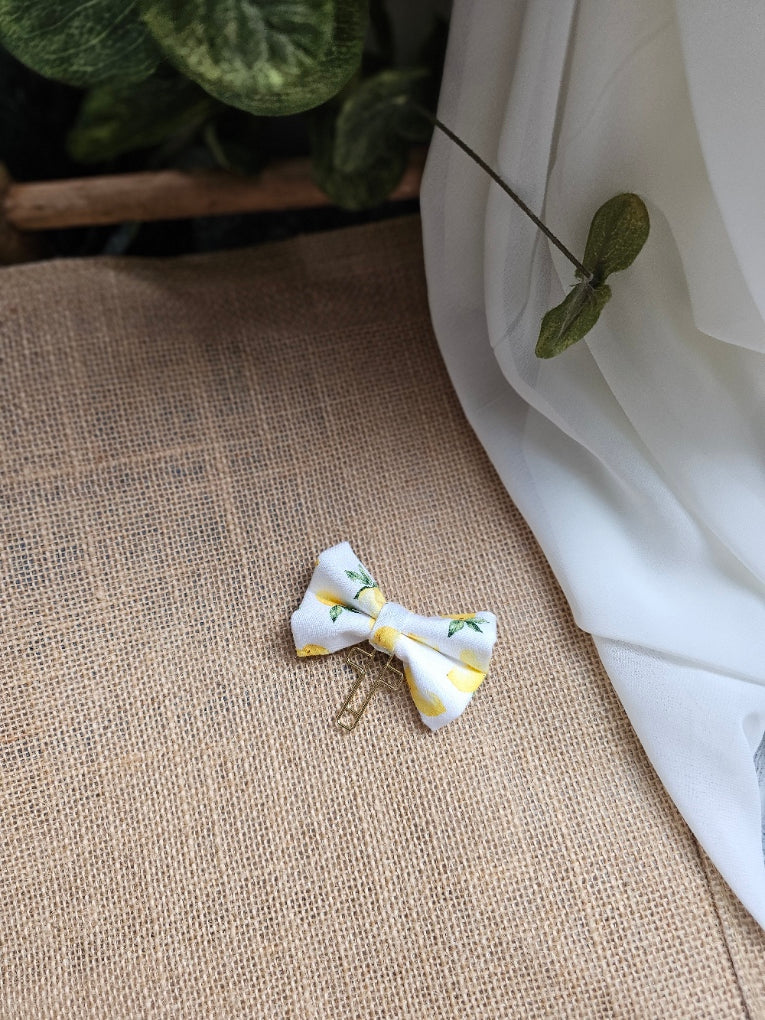 A small fabric bow bookmark with yellow lemon print, resting on a burlap surface next to white fabric and green leaves.