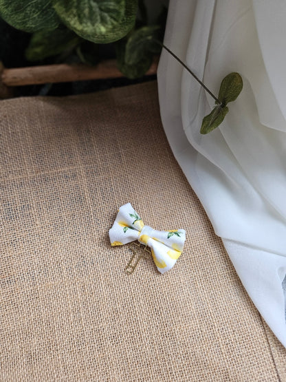 A small fabric bow bookmark with yellow lemon print, resting on a burlap surface next to white fabric and green leaves.
