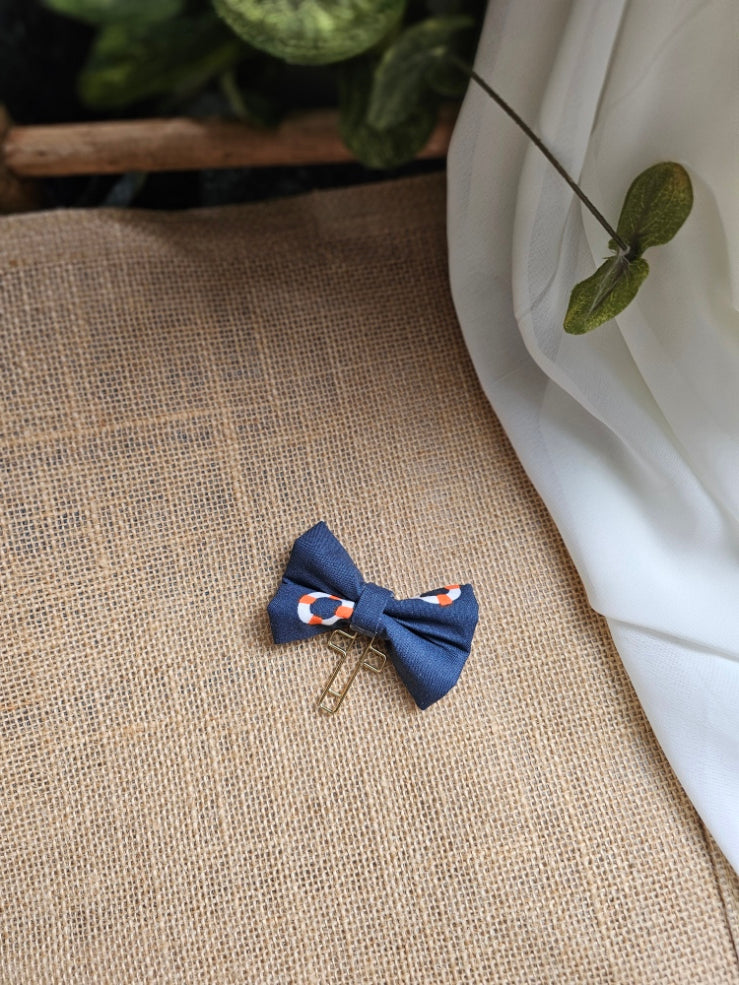 A small fabric bow bookmark with lifesaver pattern, resting on a burlap surface next to a white drape and green leaves.