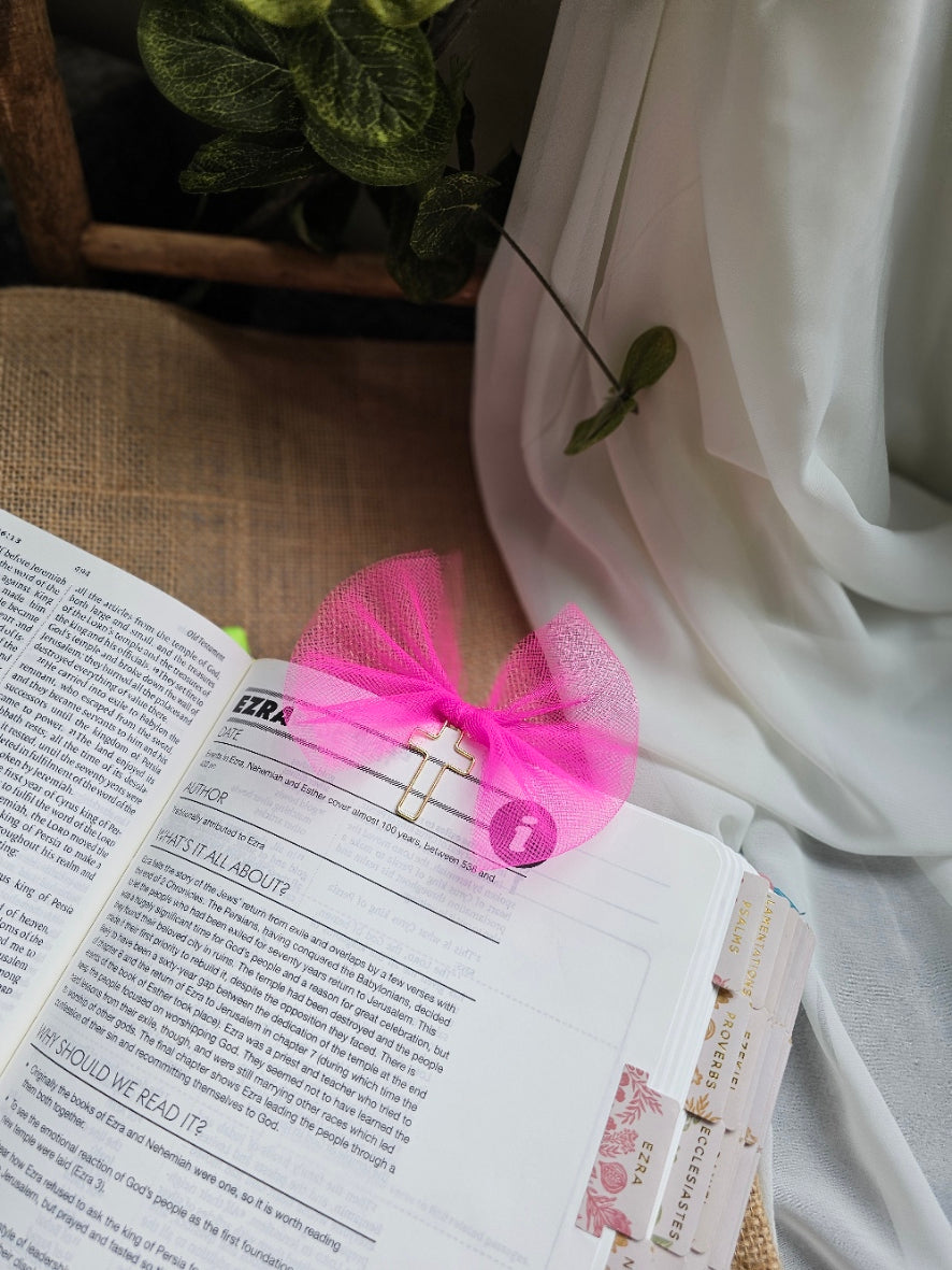 A bright pink tulle bow bookmark, attached to a metal clip and holding an open book with printed text and bookmark tabs.