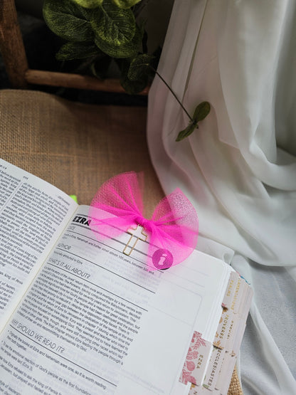 A bright pink tulle bow bookmark, attached to a metal clip and holding an open book with printed text and bookmark tabs.