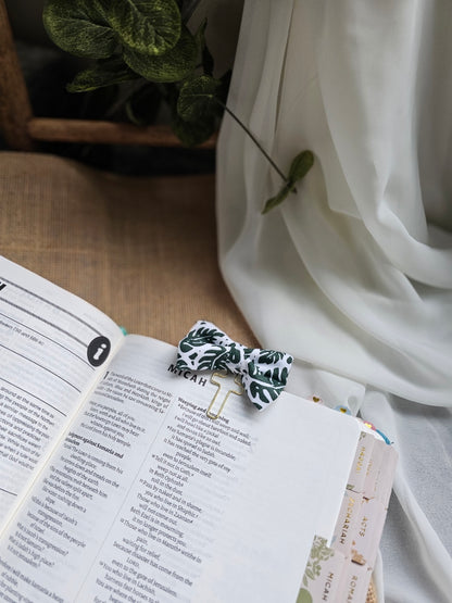 A small fabric bow bookmark with a palm pattern, holding an open book with printed text. The scene is styled with draped white fabric and a potted plant in the background.
