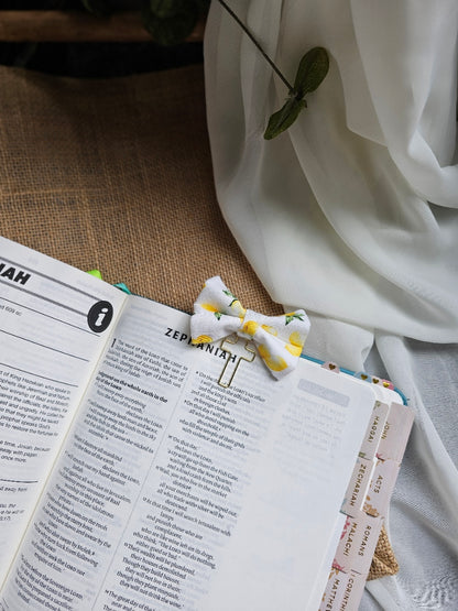 A small white bow with a yellow and green lemon pattern, held in place by a metal paperclip on an open book, styled with draped white fabric and a burlap surface.