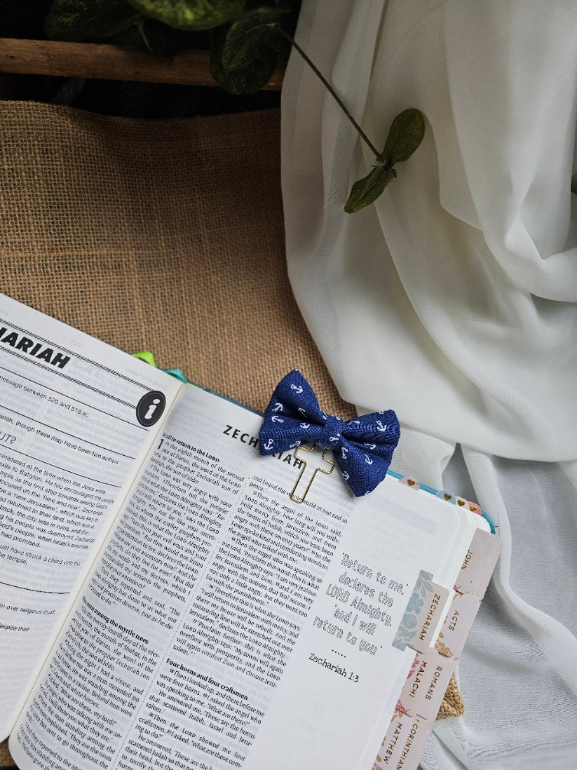 A small blue bow tie bookmark with white anchor pattern, holding open a book page. The scene includes white draped fabric, a burlap surface, and a plant leaf in the corner.