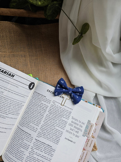 A small blue bow tie bookmark with white anchor pattern, holding open a book page. The scene includes white draped fabric, a burlap surface, and a plant leaf in the corner.