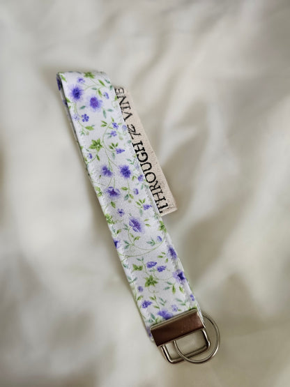 A fabric keychain strap with a white background, with purple floral pattern.