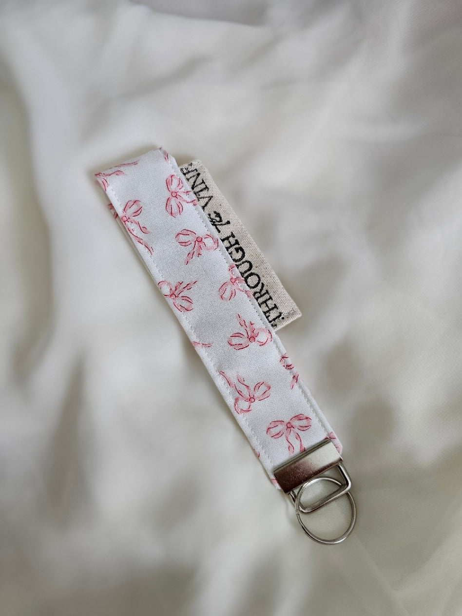 A fabric keychain strap with a white background and a pink bow pattern.