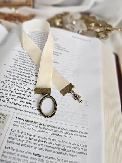 Cream velvet ribbon with brass cross and frame charms on each end sitting on top of open Bible. 